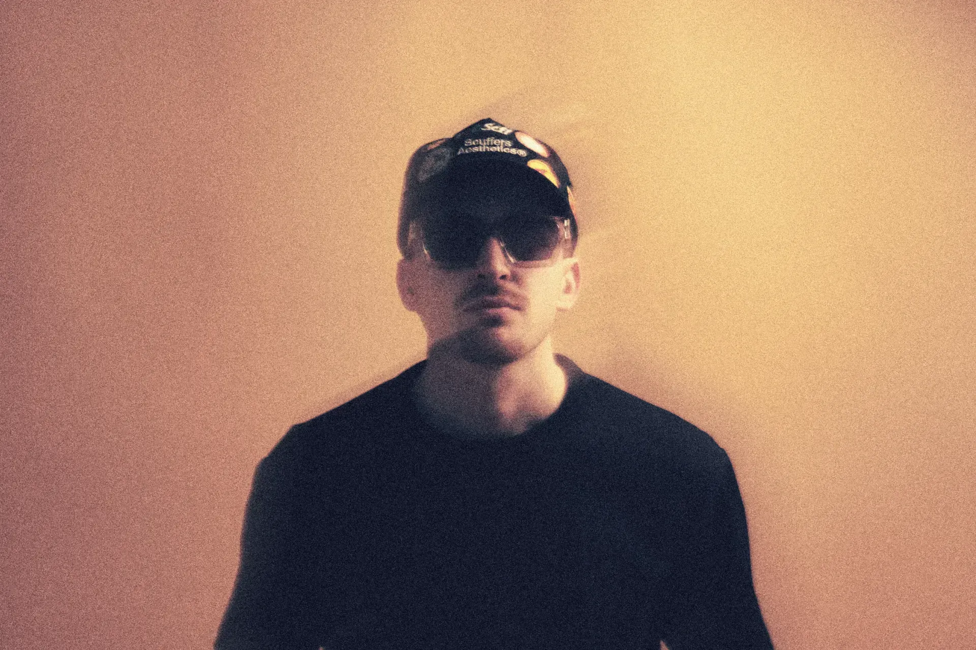 Warm sepia-toned portrait with sunglasses and cap against soft amber background