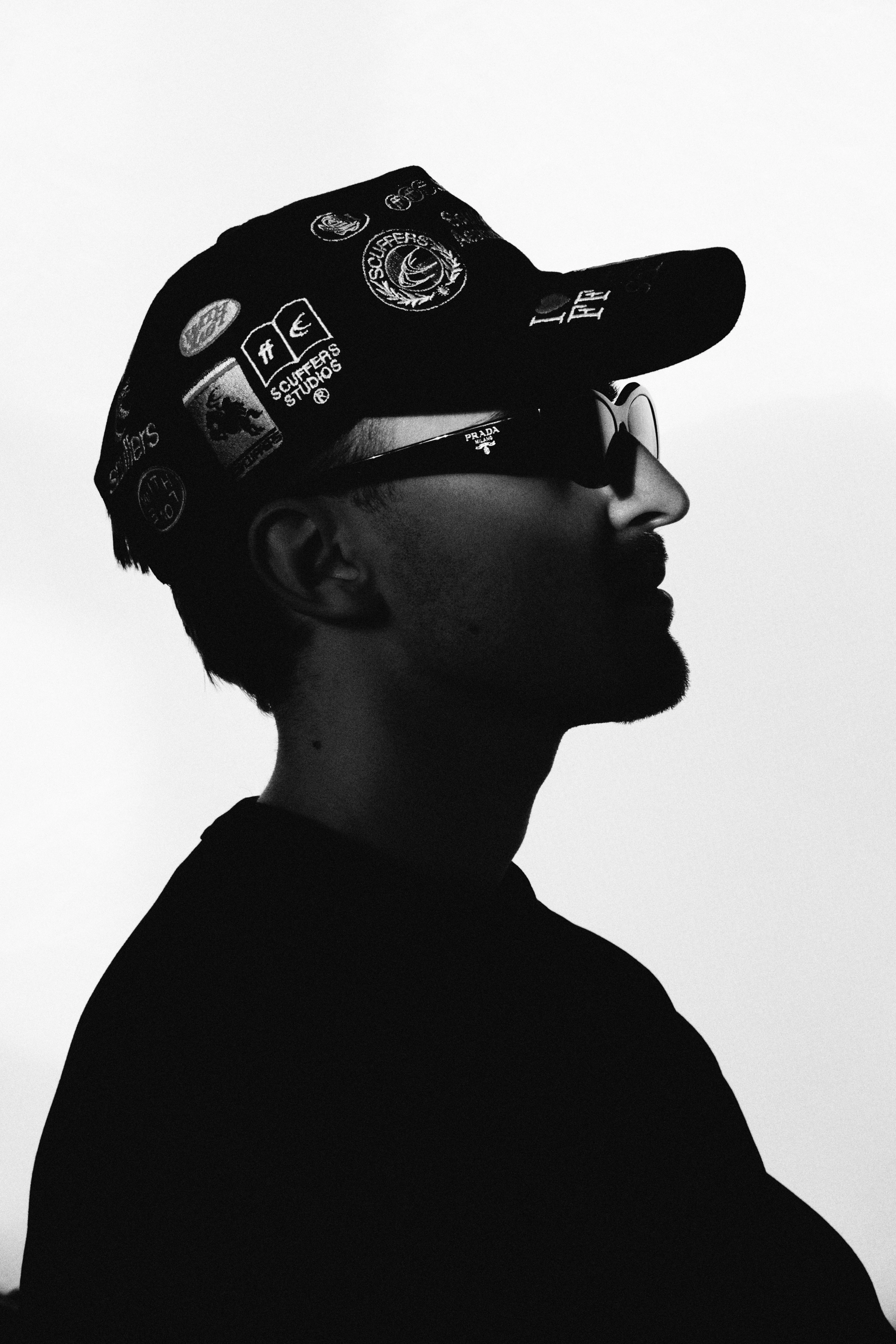 High contrast black and white side profile portrait with sticker-covered cap and sunglasses