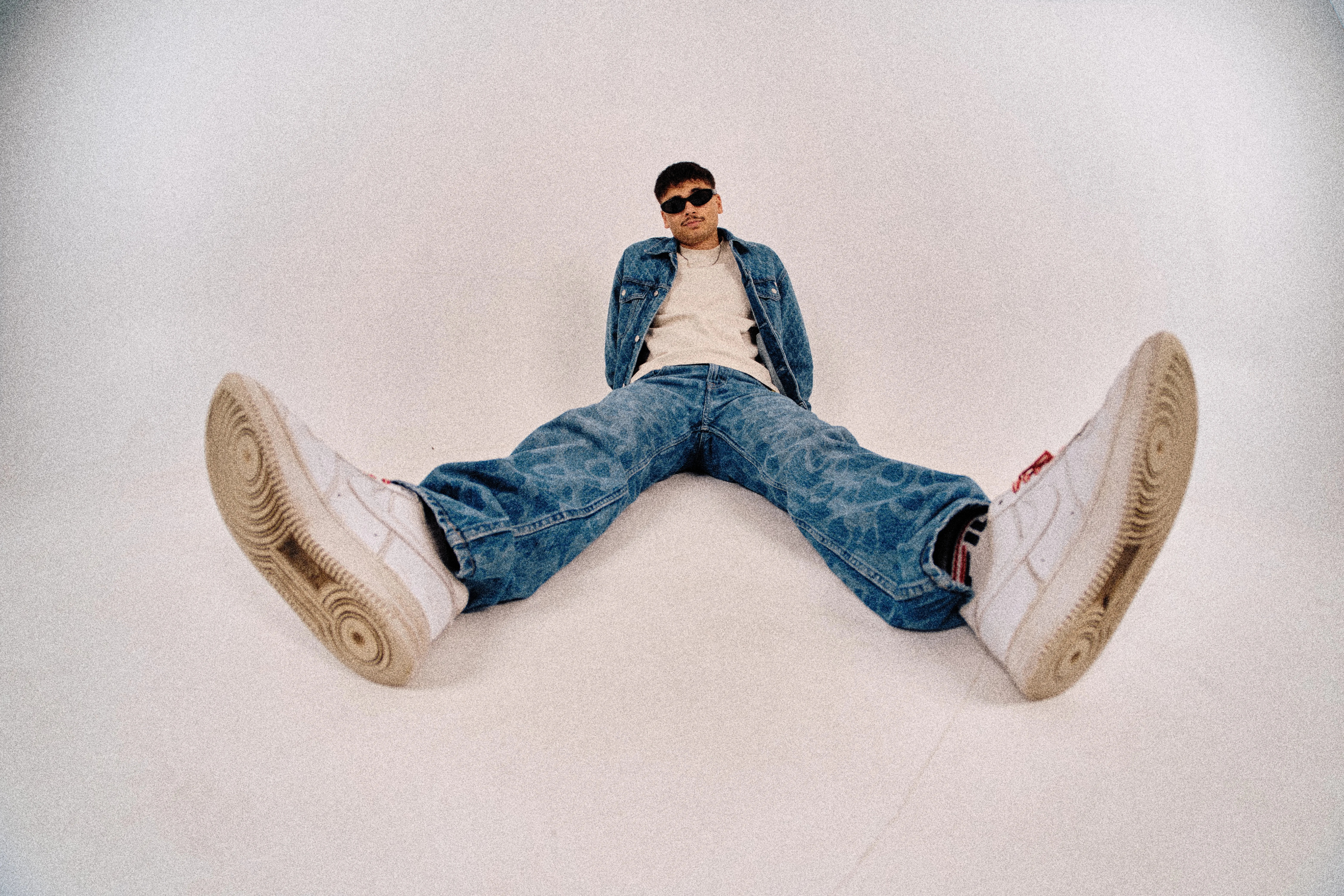 Wide-angle floor shot sitting with sneakers prominent in foreground, denim outfit and sunglasses