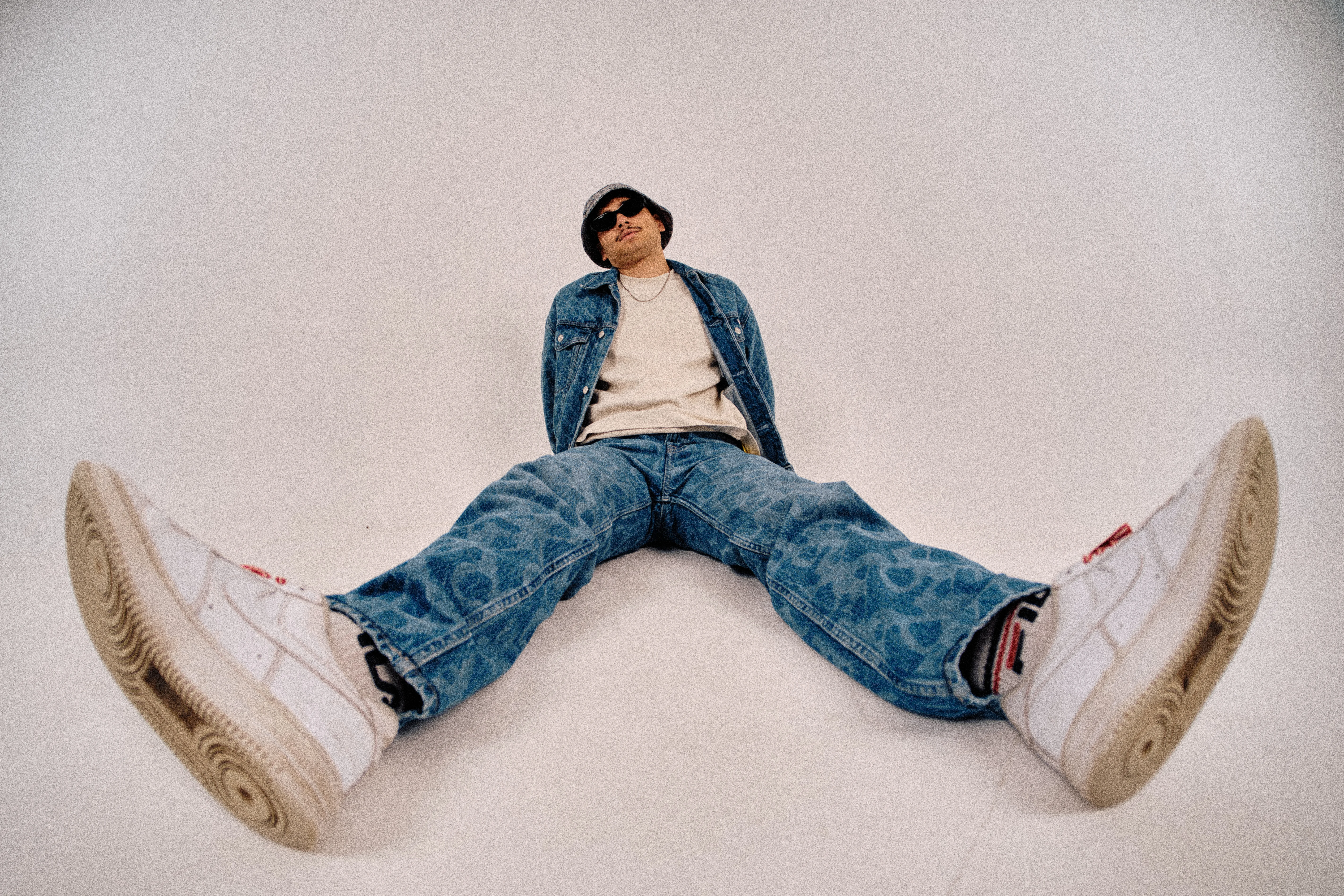 Studio portrait in denim jacket and bucket hat with sunglasses, arms crossed against grey backdrop