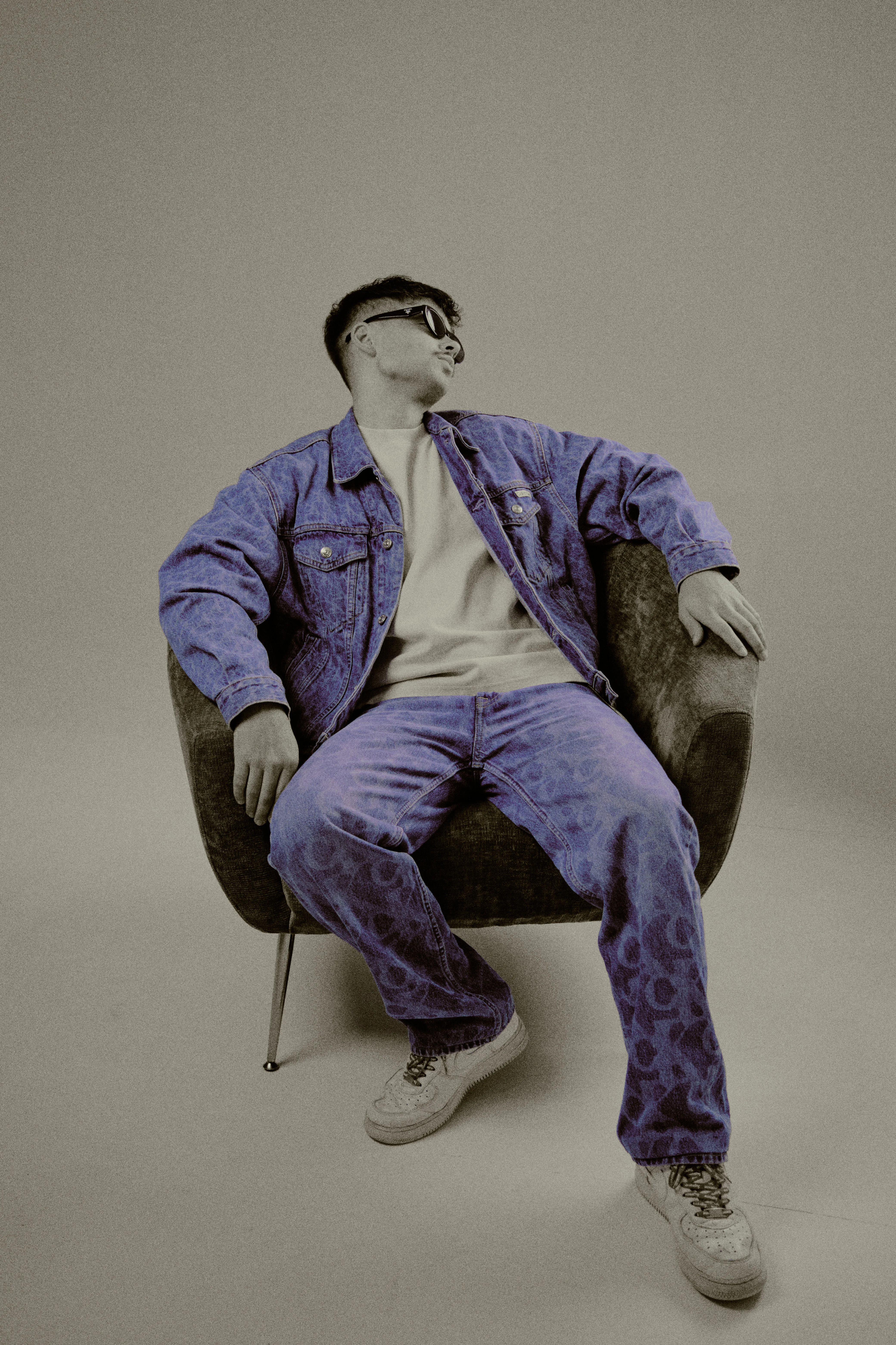 Seated in armchair wearing purple-tinted denim set with sunglasses, looking up