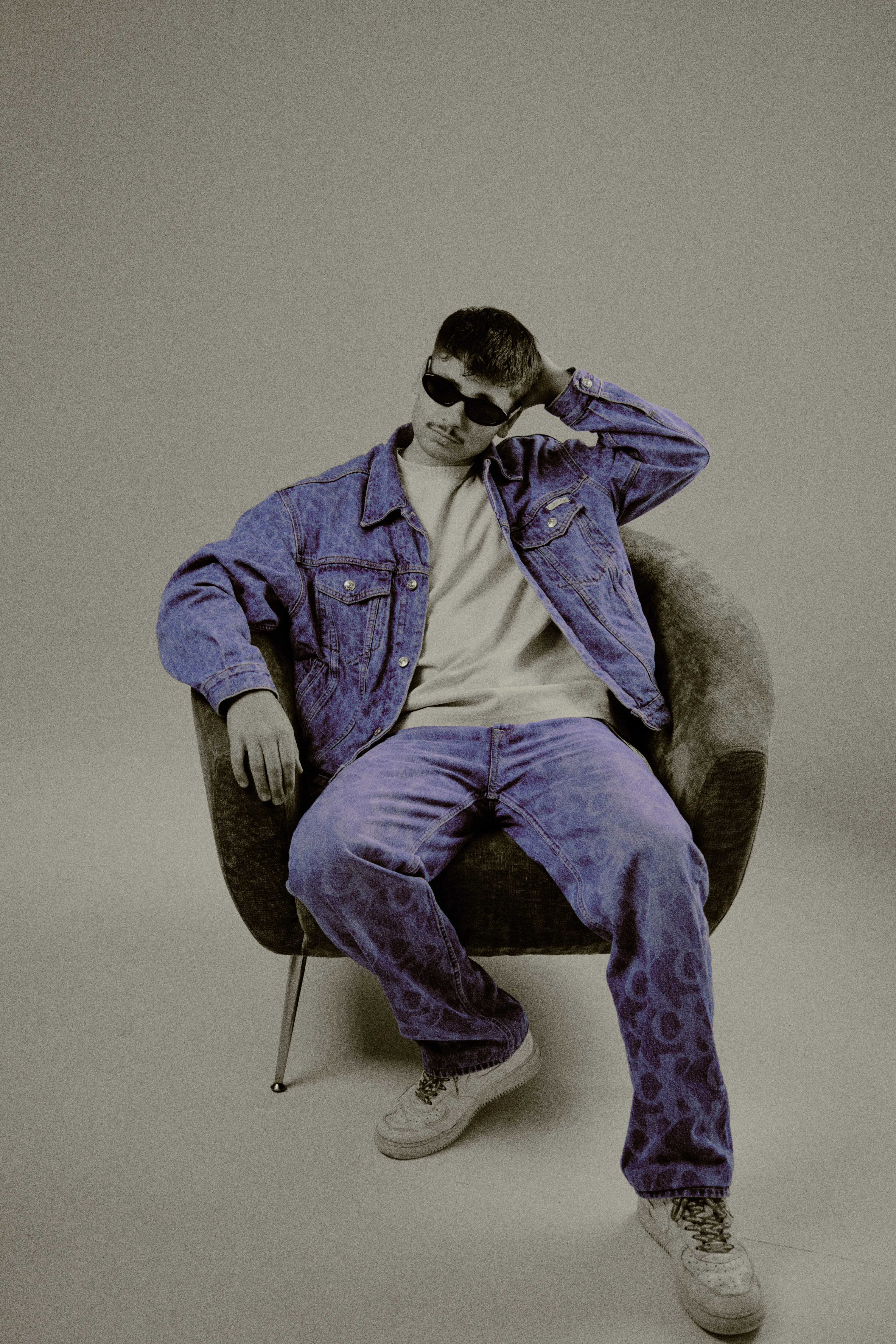 Relaxed pose in armchair with hand behind head, purple-tinted denim and sunglasses